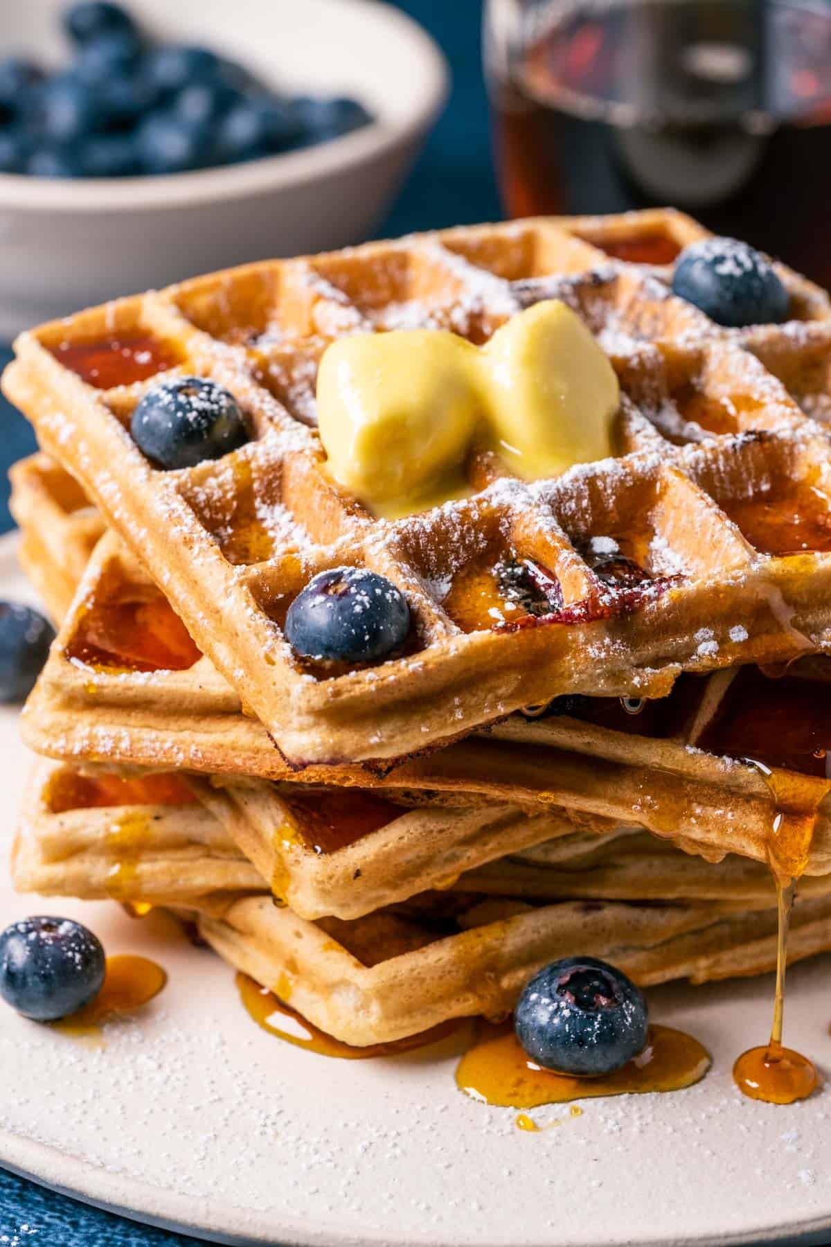 Stack of blueberry waffles topped with vegan butter, powdered sugar and syrup on a white plate.