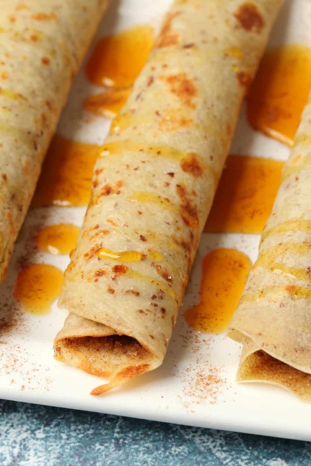 Vegan crepes rolled up on a white plate and drizzled with syrup. 